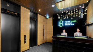 Een modern ingerichte hotellobby, met een receptiebalie, twee medewerkers achter de receptie en een digitaal bord met de naam van het hotel.