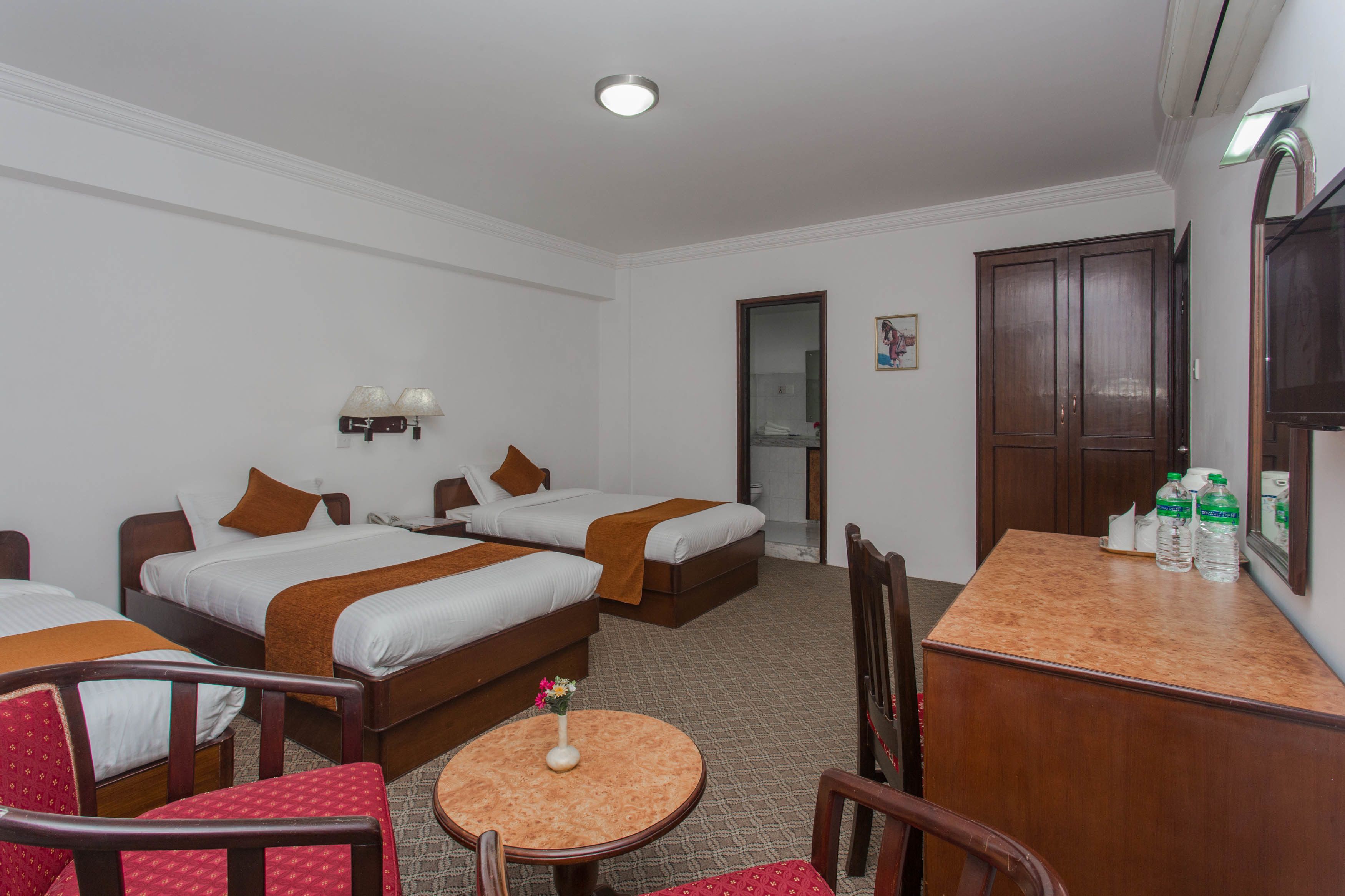 A hotel room with two single beds, a small round table with a vase, and a wooden desk with bottles.