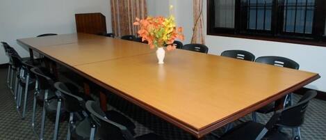 Uma sala de conferências com uma longa mesa de madeira, cadeiras pretas, um vaso branco com flores e uma janela com cortinas.