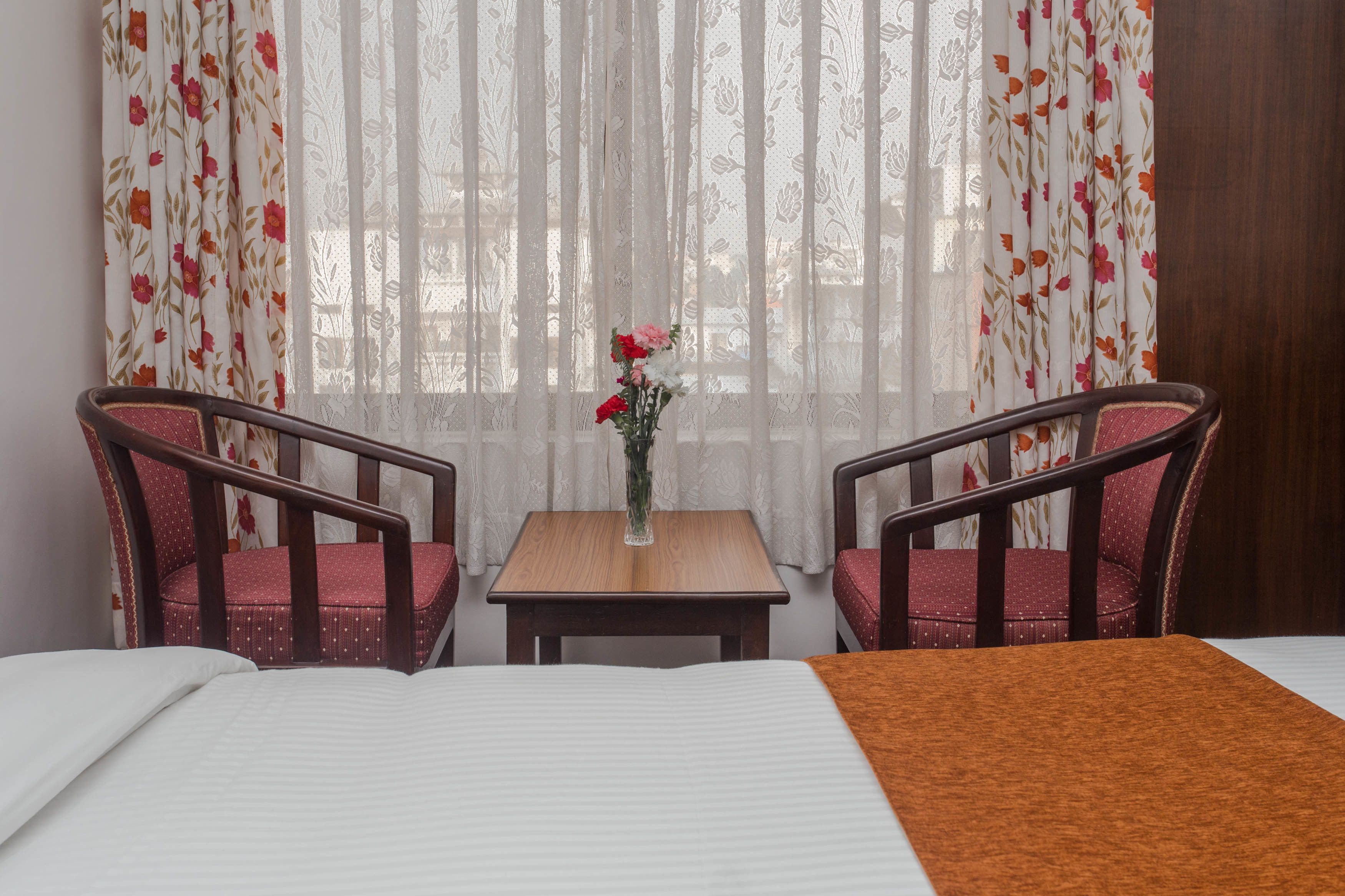 Et hotelværelse med en seng, et lille bord med en blomstervase, to stole og gardiner med et blomstermønster.