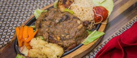 A wooden serving tray with a meat dish, vegetables, and noodles on a woven tablecloth.