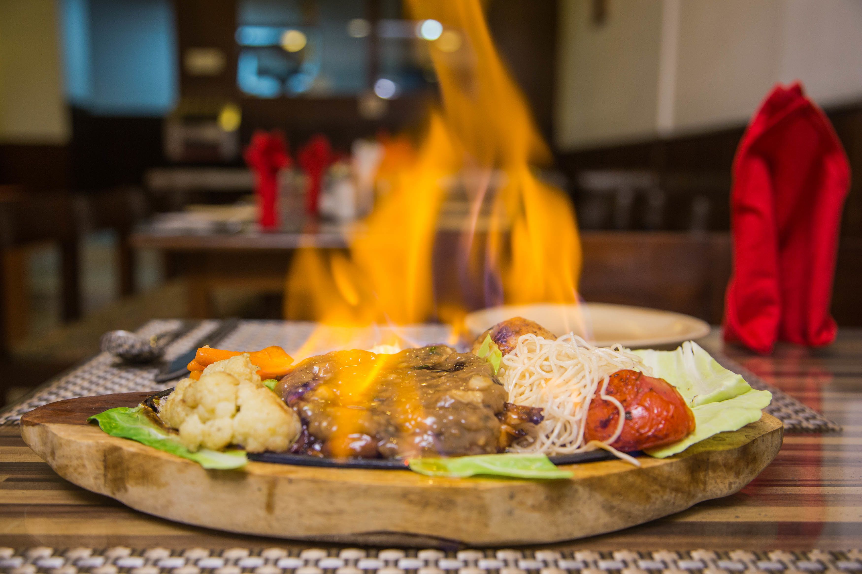 Ein Holzservierbrett mit verschiedenen Gerichten, darunter ein Fleischpastetchen, Gemüse und Nudeln, auf einem Tisch, im Hintergrund brennt ein Feuer.