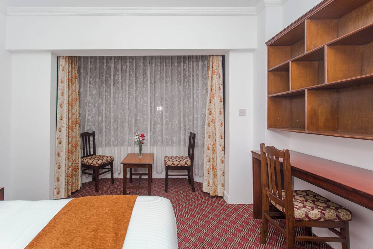Hotellihuone, jossa on sänky, työpöytä, kaksi tuolia, pieni pöytä maljakolla ja suuri puinen hylly.