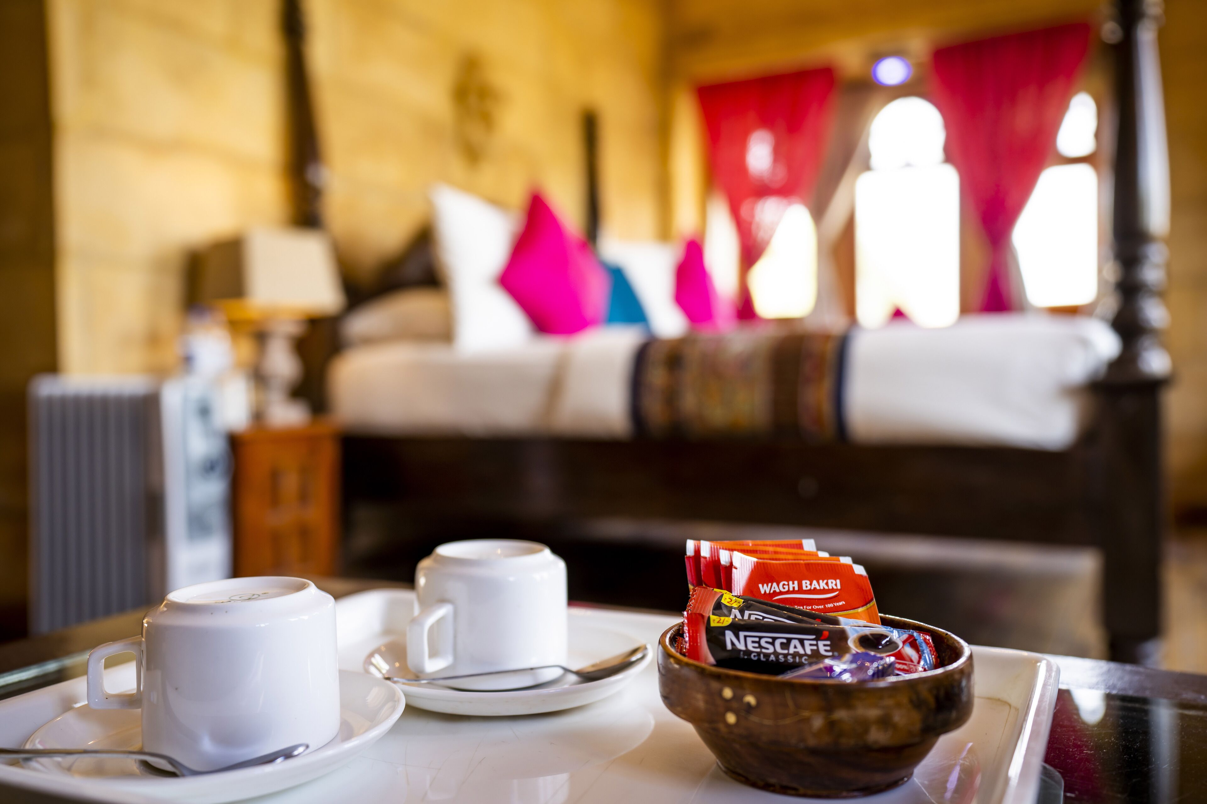 Un vassoio della colazione con due tazze bianche, una ciotola di Nescafe Classic e un cucchiaio su un tavolo in una stanza con un letto e tende rosse sullo sfondo.