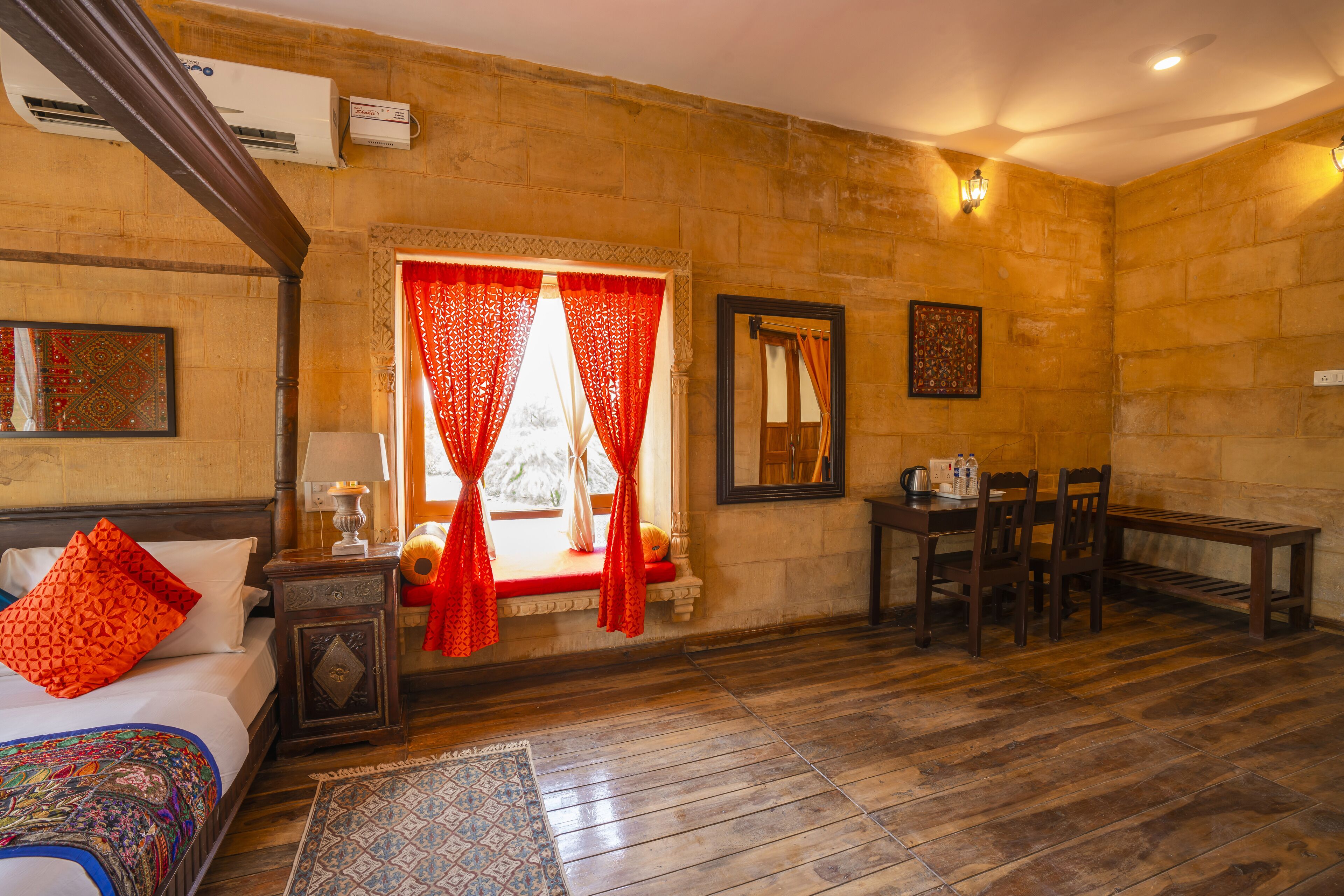A hotel room with a wooden floor, a bed with a colorful blanket, a small table with a lamp, a window with red curtains, and a dining area with chairs and a table.