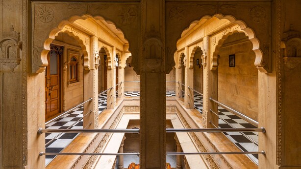 Un pasillo con arcos ornamentales y suelo a cuadros, que conduce a una puerta al final.