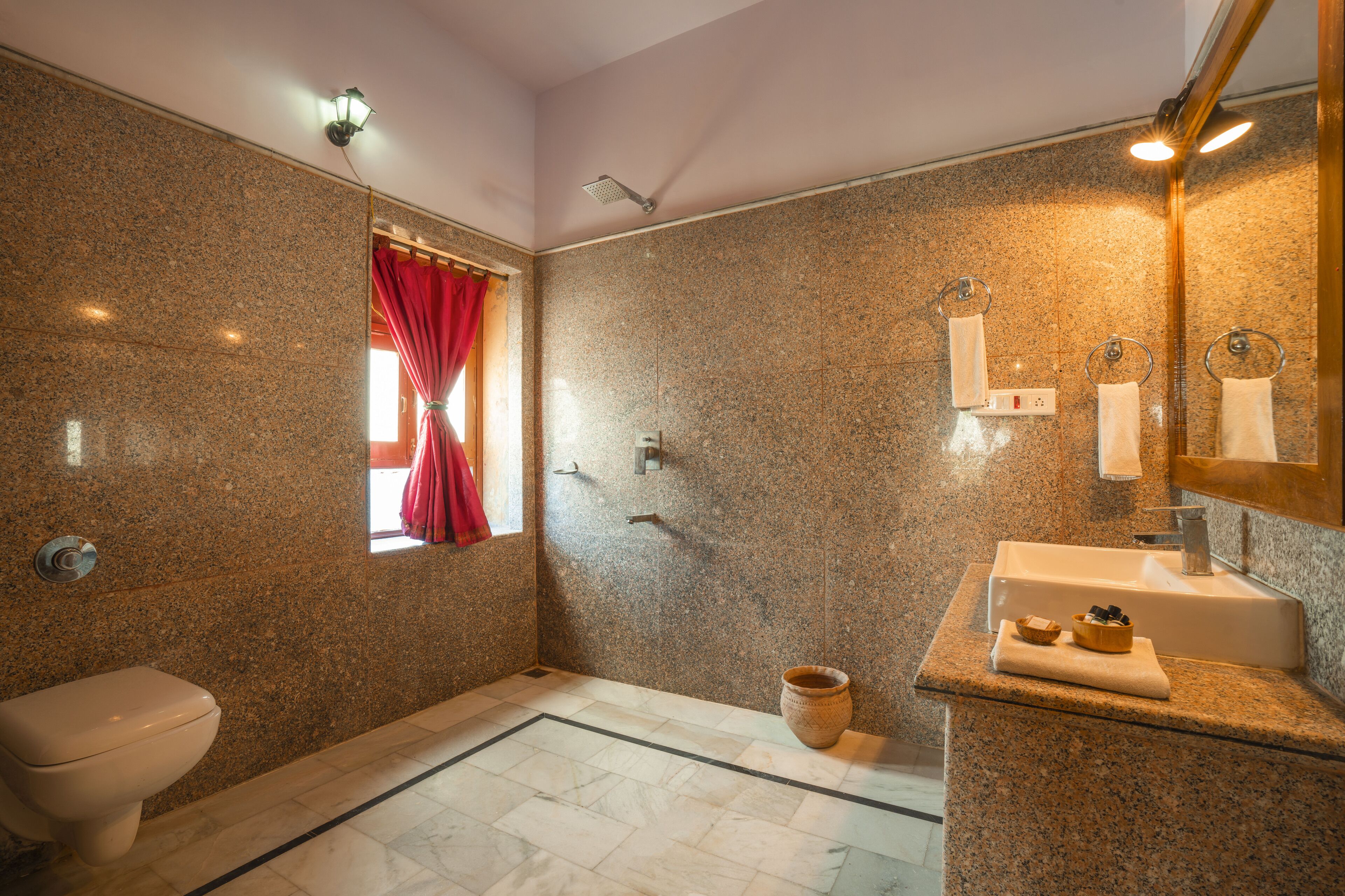 A bathroom with a granite wall, a shower, a toilet, a sink, and a window with a red curtain.