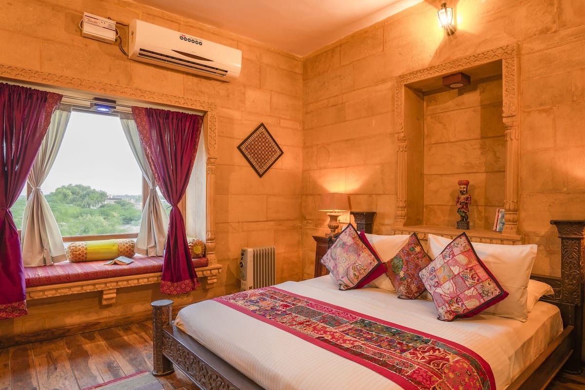 A bedroom with a large bed, a window with curtains, an air conditioner, and a view of greenery outside.