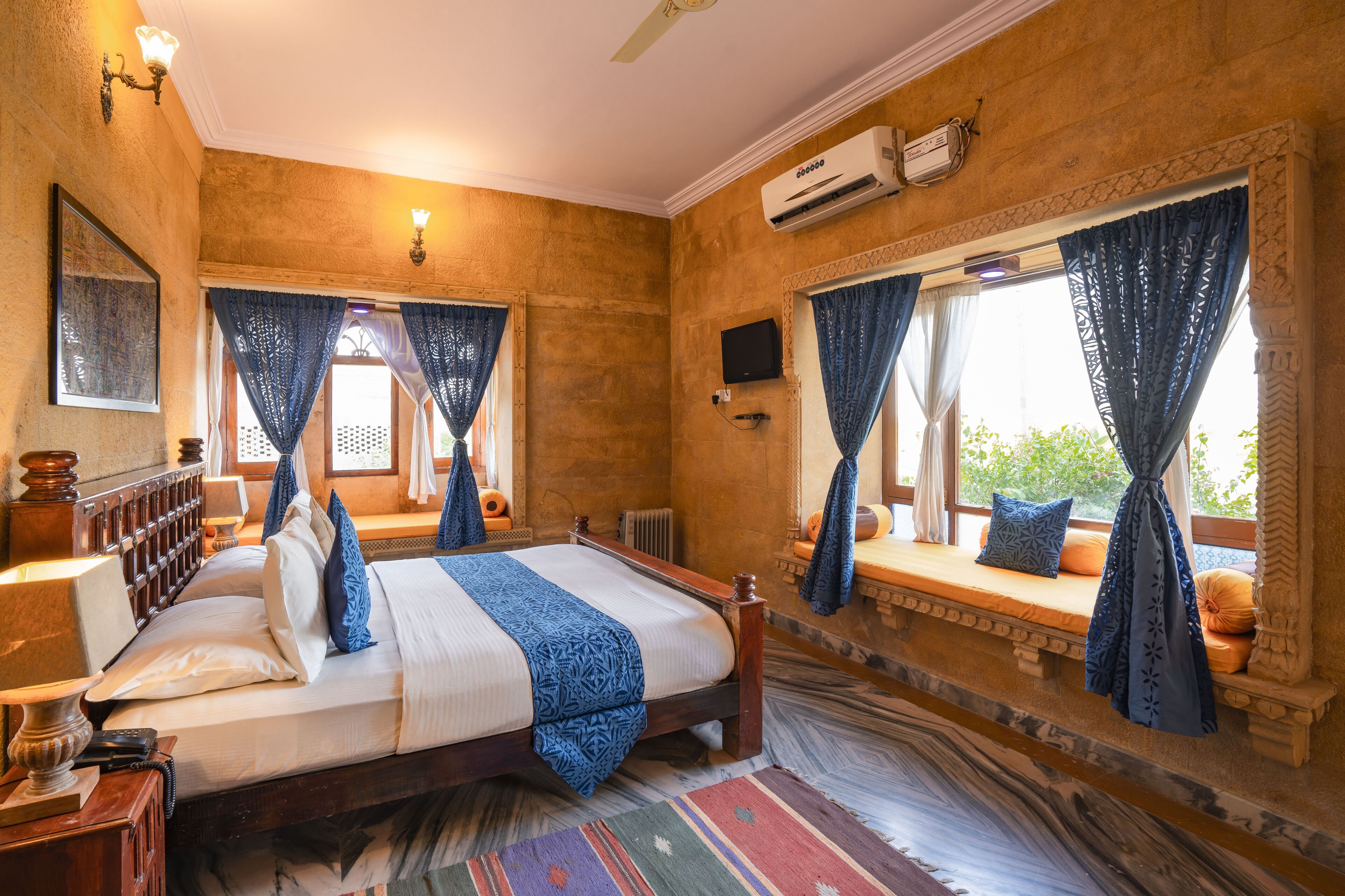 A bedroom with a bed, a bedside table, a lamp, a window with curtains, and a wall-mounted air conditioner.