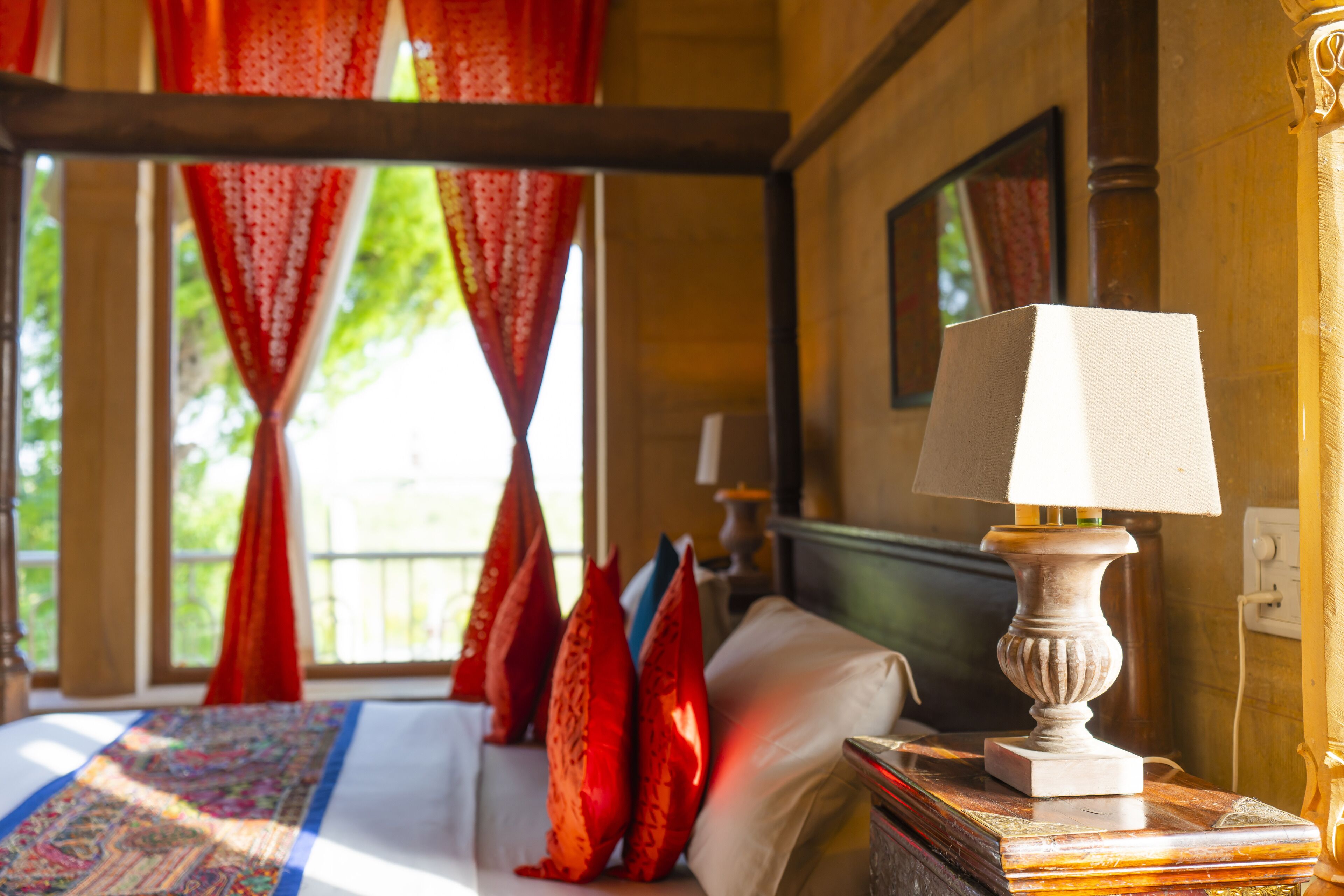 A room with a bed, red pillows, a white lamp, and a window with red curtains.