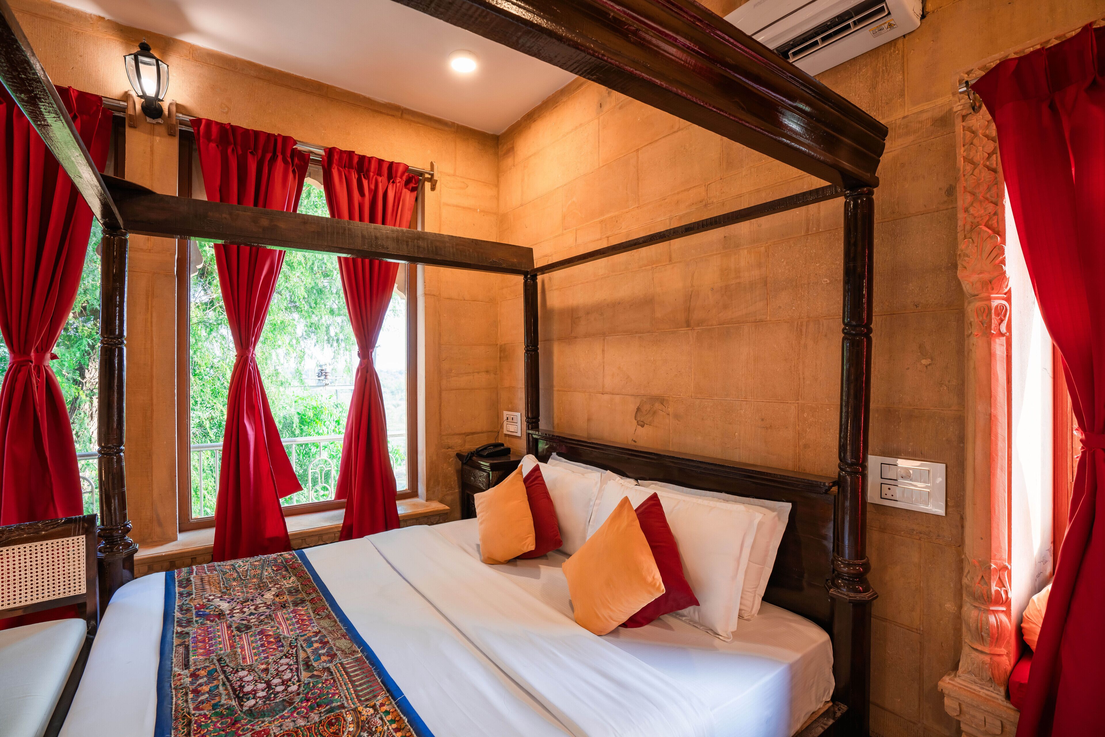 A four-poster bed with red curtains, a patterned rug, and a view of greenery outside.