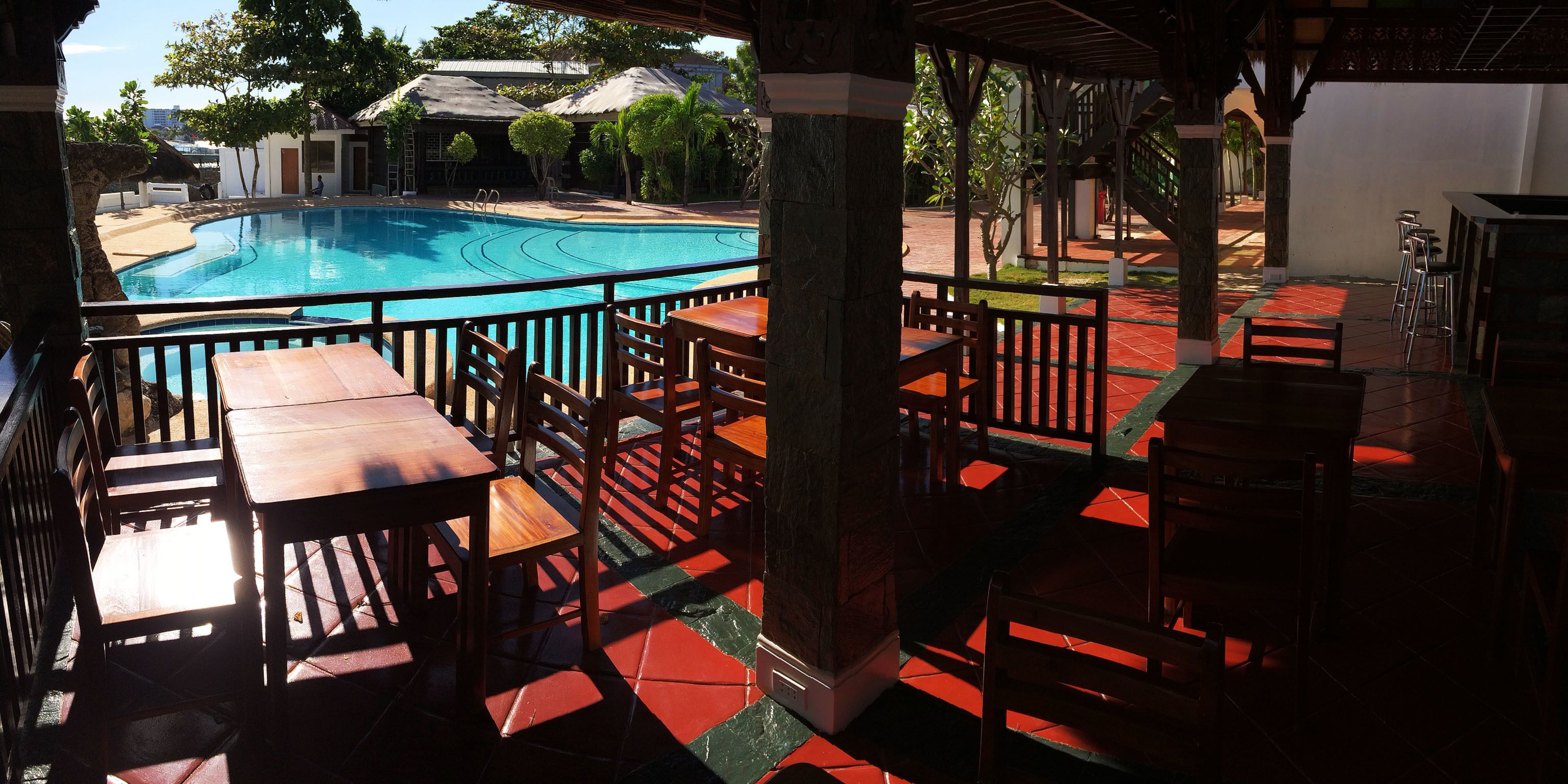 Un espace aménagé au bord de la piscine, avec des tables et des chaises en bois, une piscine et un coin salon couvert offrant une vue sur la piscine.