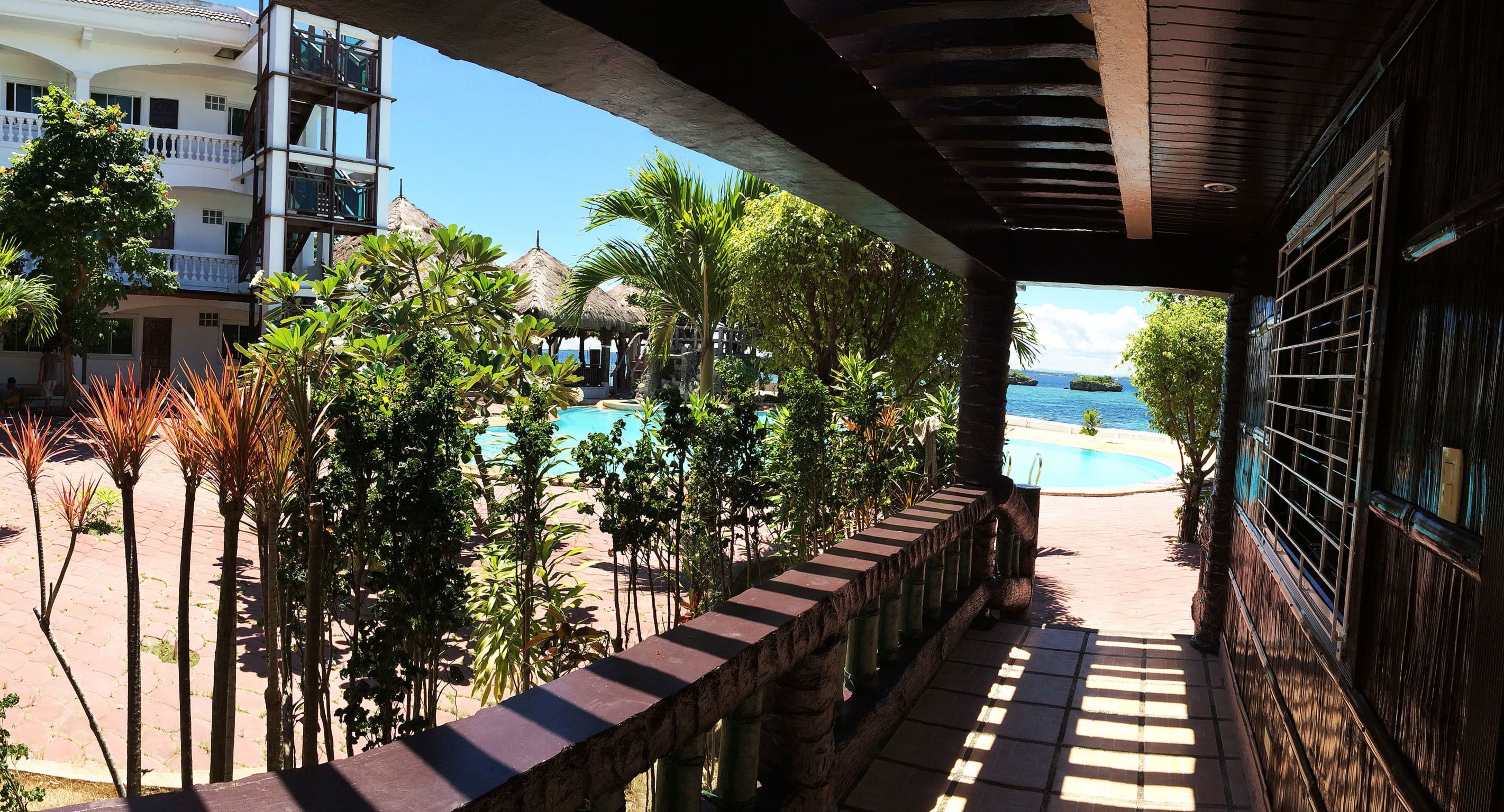 Vista de un área de piscina con terraza de madera y plantas tropicales, que da a una playa con una vista despejada del océano.