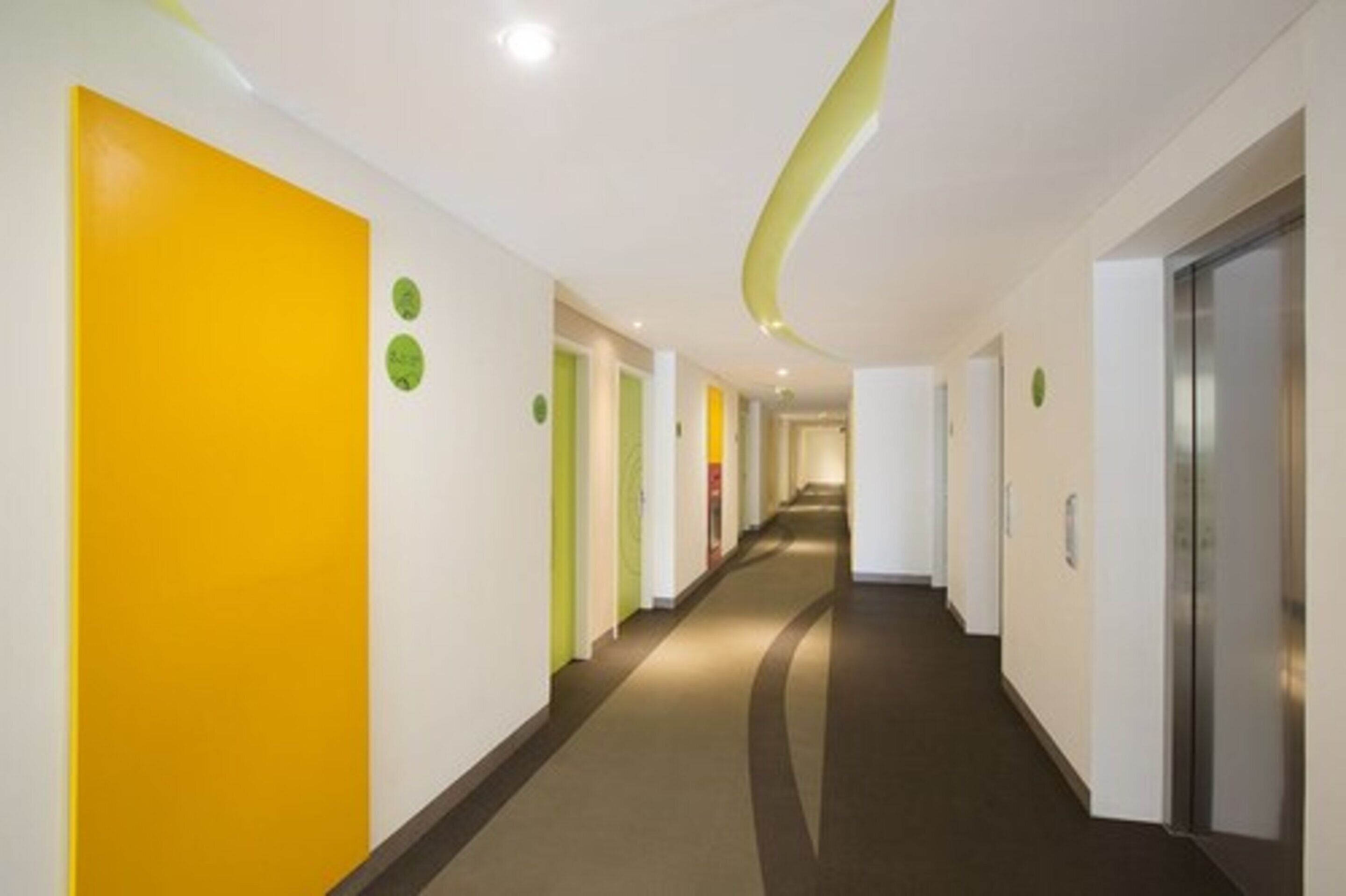 Un couloir d’hôtel moderne avec un plafond incurvé, des murs jaune vif et verts, et un sol recouvert de moquette.