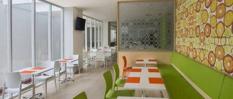 Un comedor moderno con mesas blancas y naranja, sillas verdes y una pared con estampado.