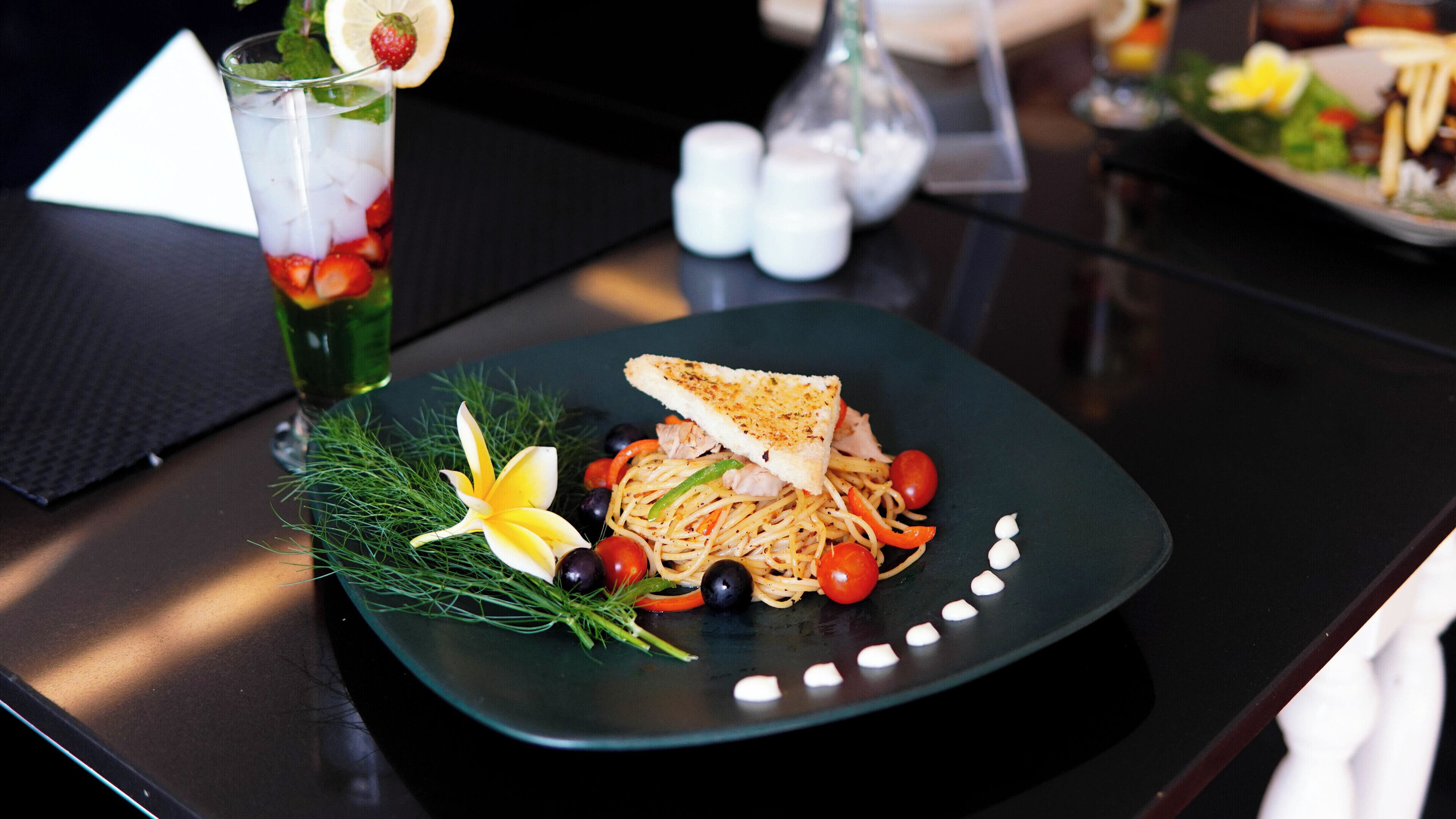 Un plat de spaghetti amb verdures i una llesca de pa torrat, decorat amb una flor i herbes, damunt d'una taula fosca.