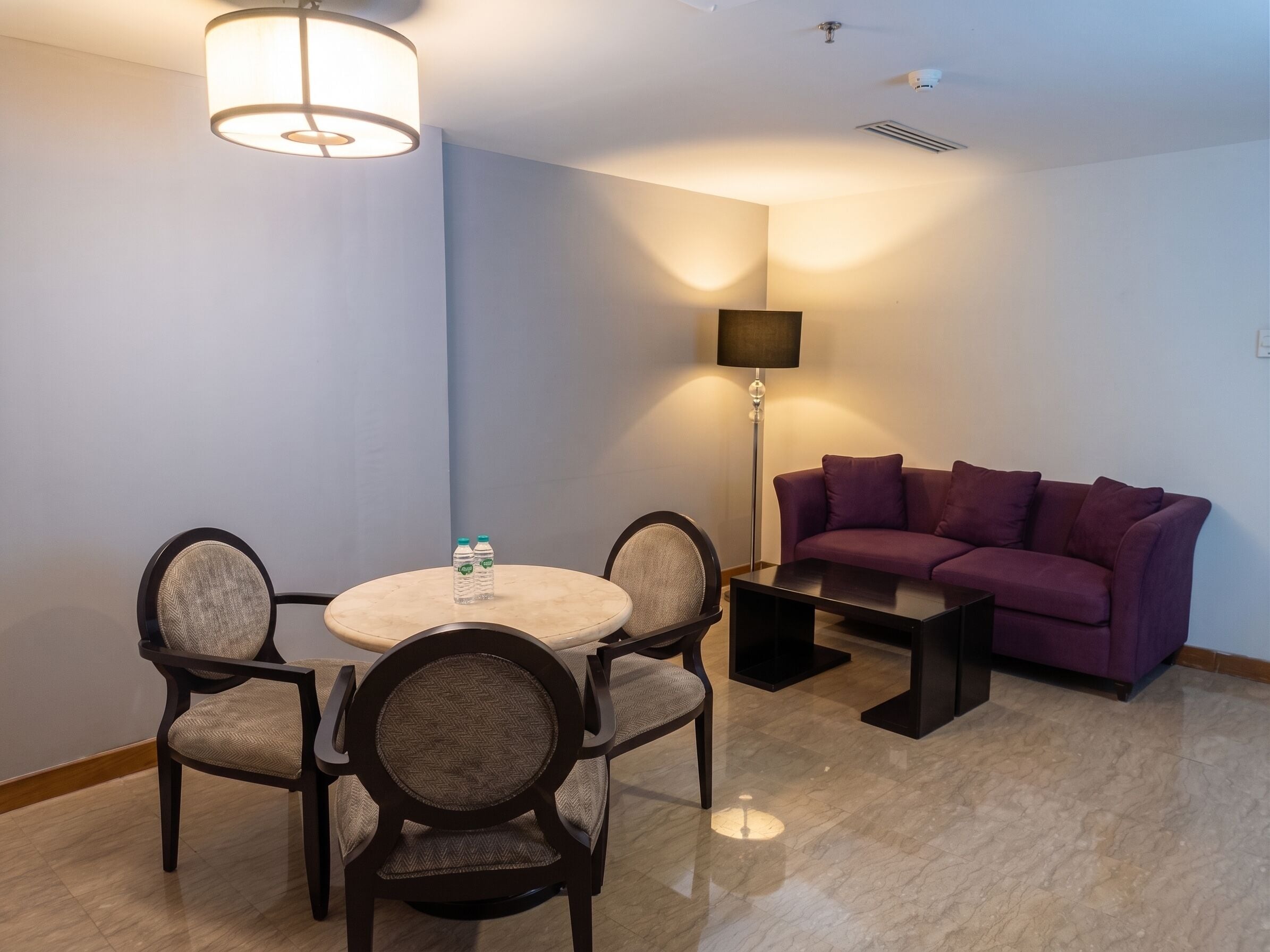 Una habitación de hotel con una mesa redonda de comedor, cuatro sillas, un sofá morado, una lámpara de pie y una botella de agua sobre la mesa.