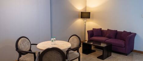Una habitación de hotel con una mesa redonda de comedor, cuatro sillas, un sofá morado, una lámpara de pie y una botella de agua sobre la mesa.
