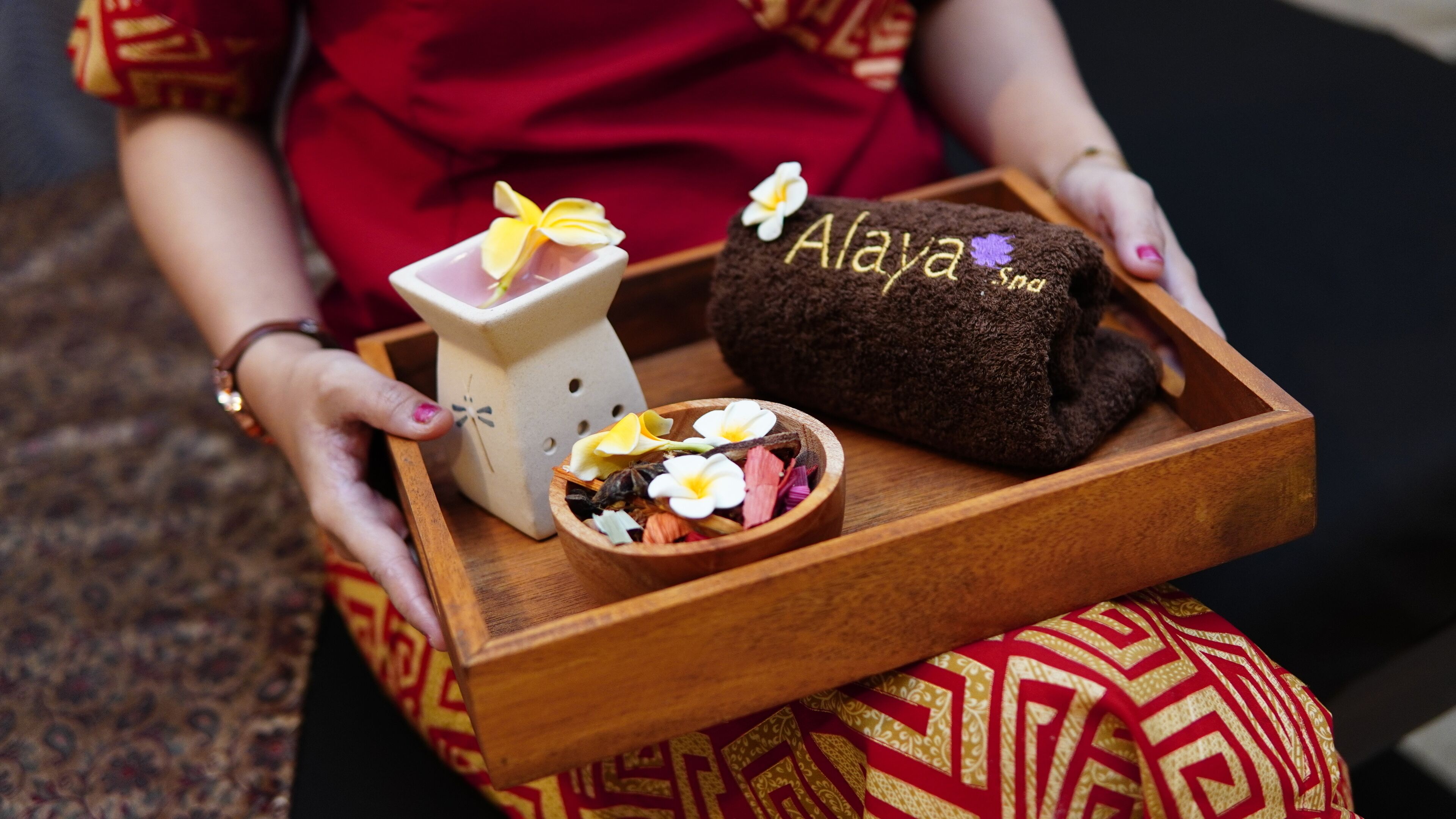 Una persona sostiene una bandeja de madera con una maceta de cerámica, un cuenco de flores y un paño marrón con el bordado 'Alaya Spa'.