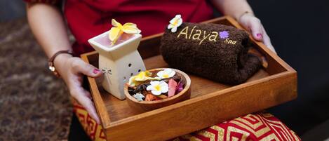 Una persona sostiene una bandeja de madera con una maceta de cerámica, un cuenco de flores y un paño marrón con el bordado 'Alaya Spa'.