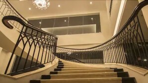 Una gran escalera con barandales negros y un candelabro, que conduce a un segundo piso con ventanas amplias.