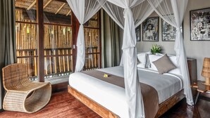 Un dormitorio con cama con dosel, una silla de mimbre y un ventanal con persianas de bambú.