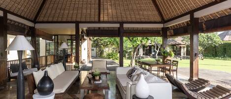A covered outdoor seating area with a thatched roof, cushioned seating, and a dining table.