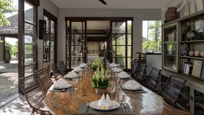 Un comedor con una larga mesa de madera dispuesta para una comida, rodeado de sillas de mimbre, un ventilador de techo y amplios ventanales que dejan entrar luz natural.