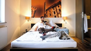 A hotel room with a bed, two bedside lamps, a wall-mounted painting, and a door with a towel hanging on it. - Pfefferbett Hostel Berlin (Berlin)