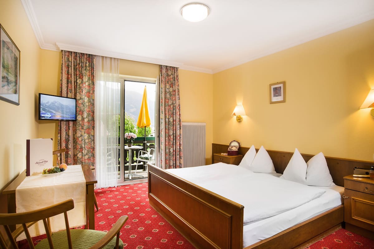 Ein Hotelzimmer mit einem großen Bett, einem Schreibtisch mit Computer, einem Sessel und Blick ins Freie durch eine Schiebetür.