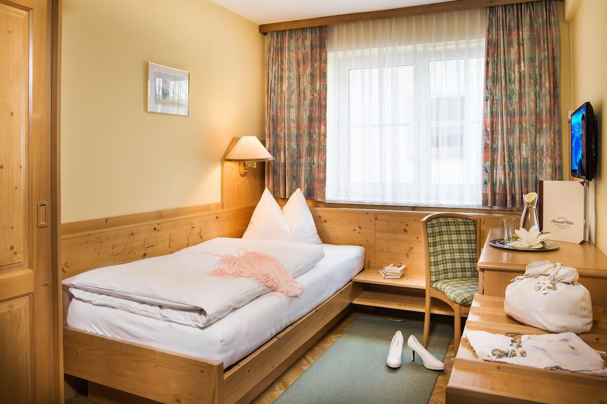 Ein Hotelzimmer mit einem hölzernen Bett, einem Schreibtisch mit Computer, einem Stuhl und einem Fenster mit Vorhängen.