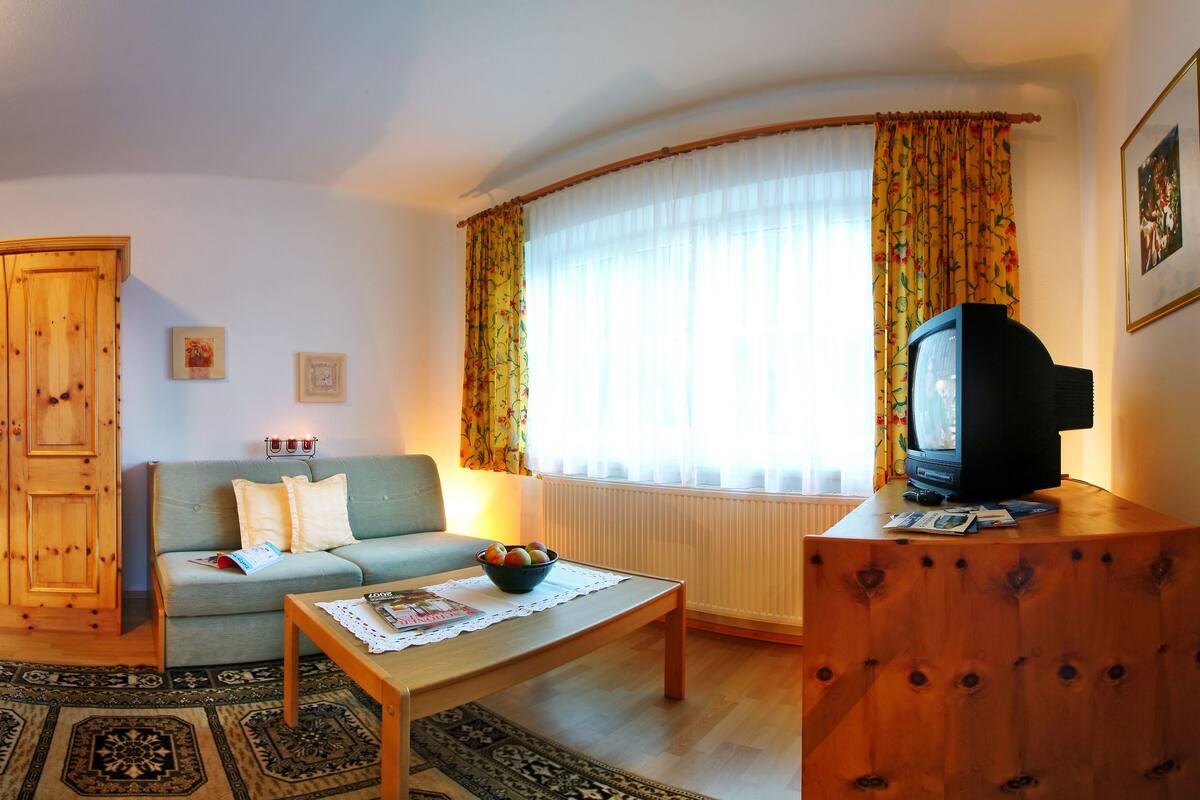 Ein gemütliches Wohnzimmer mit einer Couch, einem Sessel, einem Holzschrank, einem Fernseher und einem kleinen Tisch mit einer Obstschale.