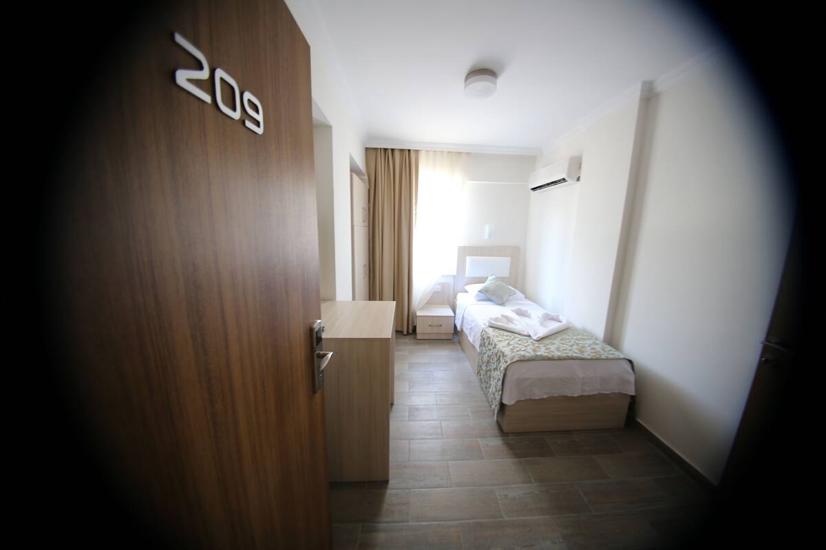 Ein Hotelzimmer mit einer Holztür mit der Aufschrift "209", einem Bett mit weißem Überwurf, einem Nachttisch und einem Fenster mit Vorhängen.