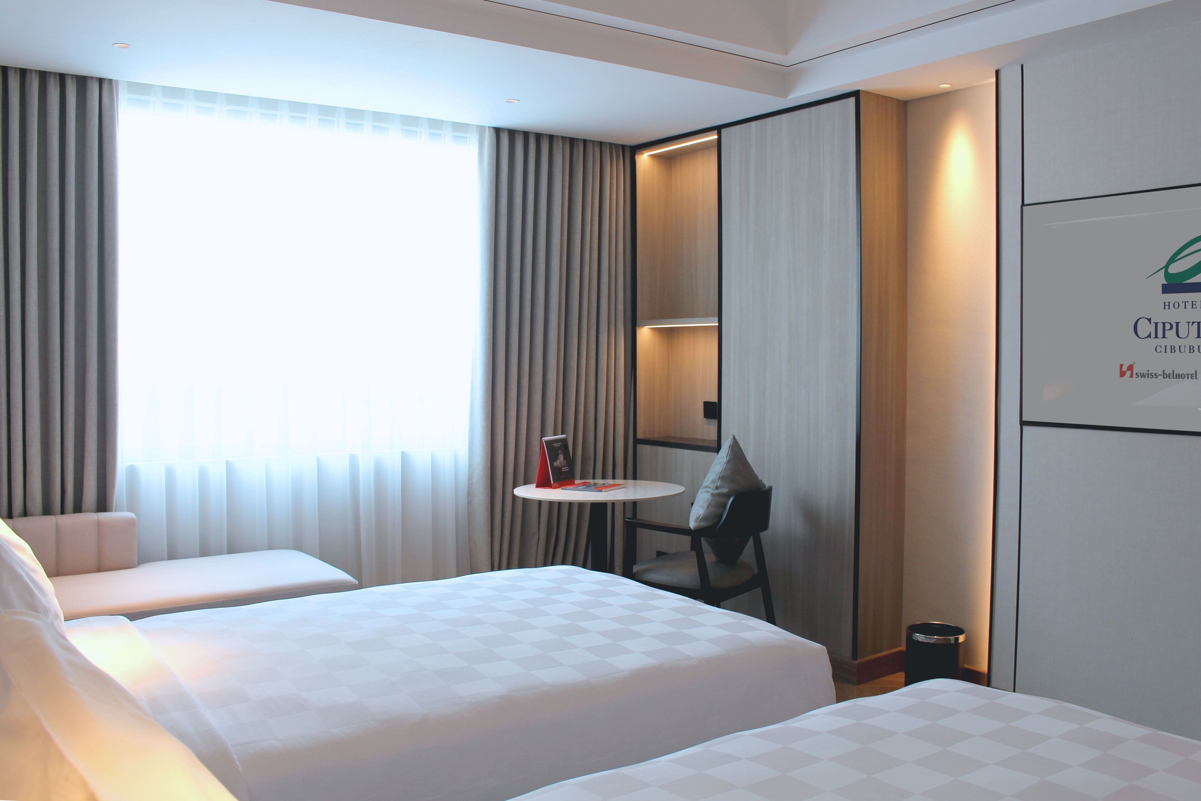 A hotel room with two beds, a small table, a chair, and a window with curtains.
