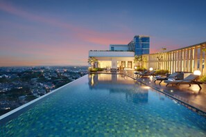 Piscina infinita en la azotea con sillones de descanso y vistas a la ciudad al atardecer.