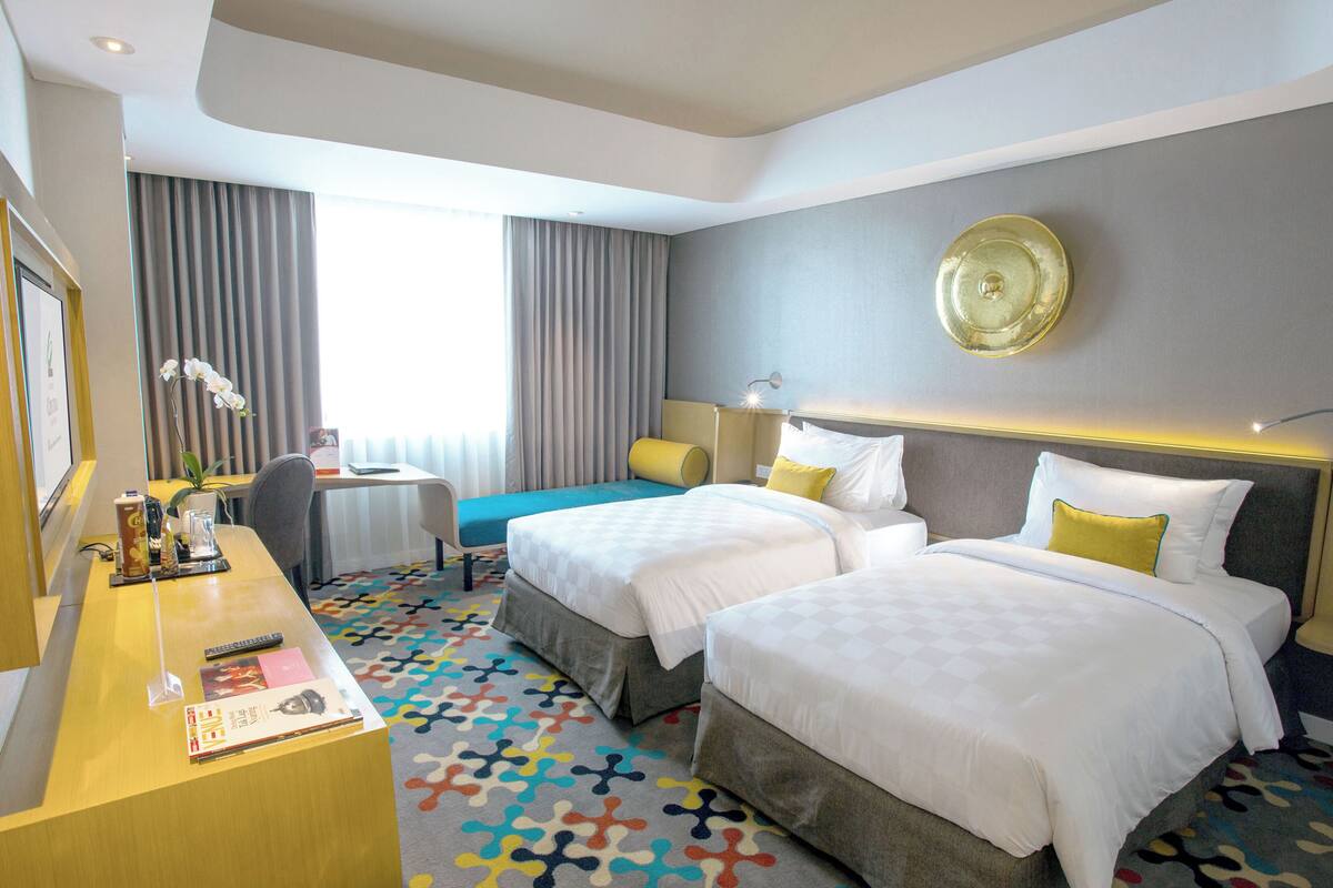 A hotel room with two beds, a desk with a chair, a small table, and a window with curtains.