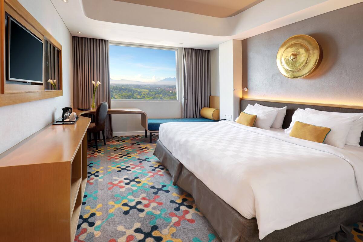 A hotel room with a large bed, a desk with a chair, a TV, and a view of the mountains.