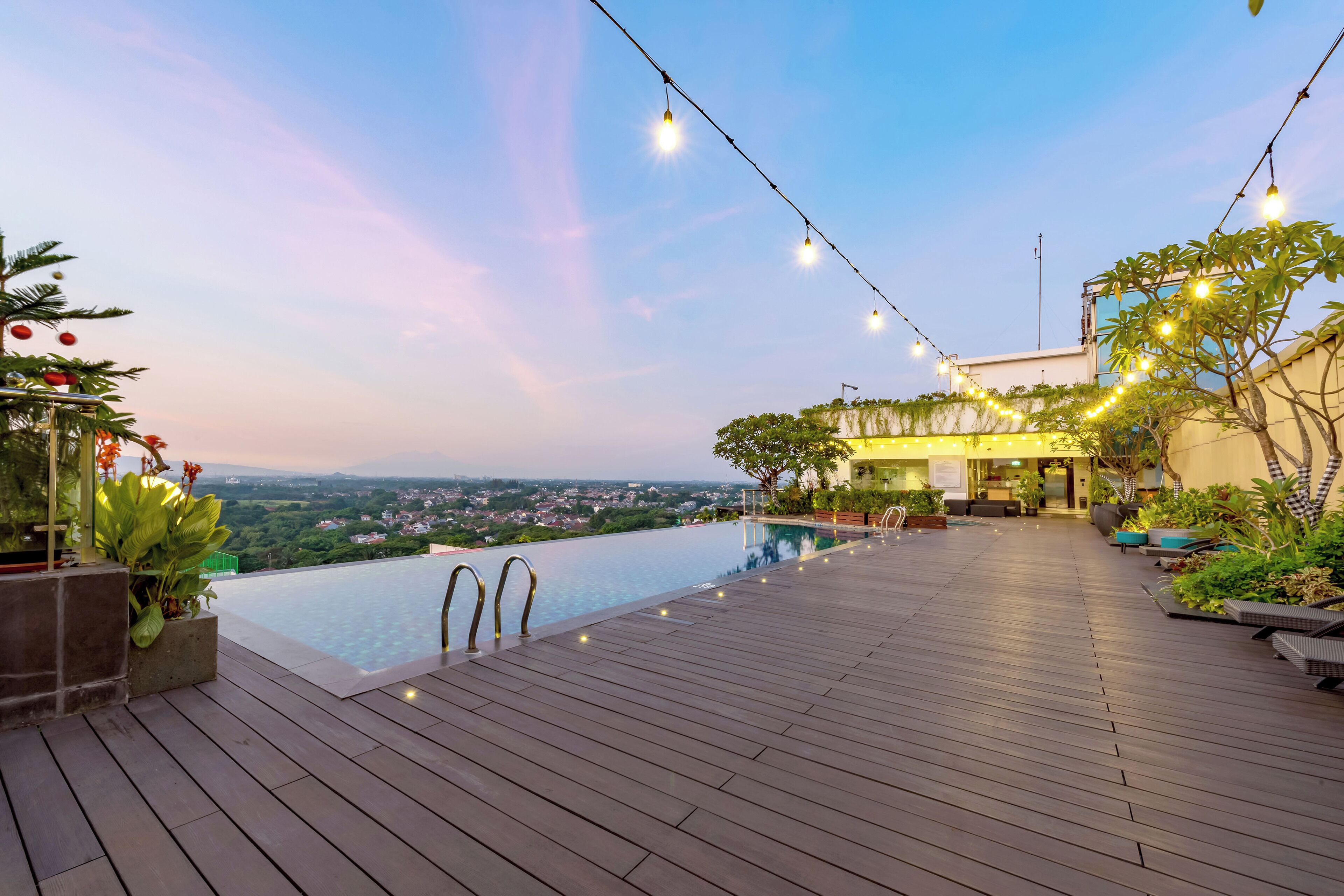 A rooftop infinity pool with wooden decking, string lights, and a view of the cityscape.