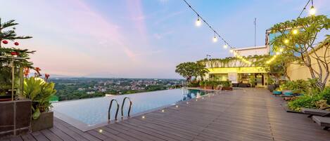 A rooftop infinity pool with wooden decking, string lights, and a view of the cityscape.