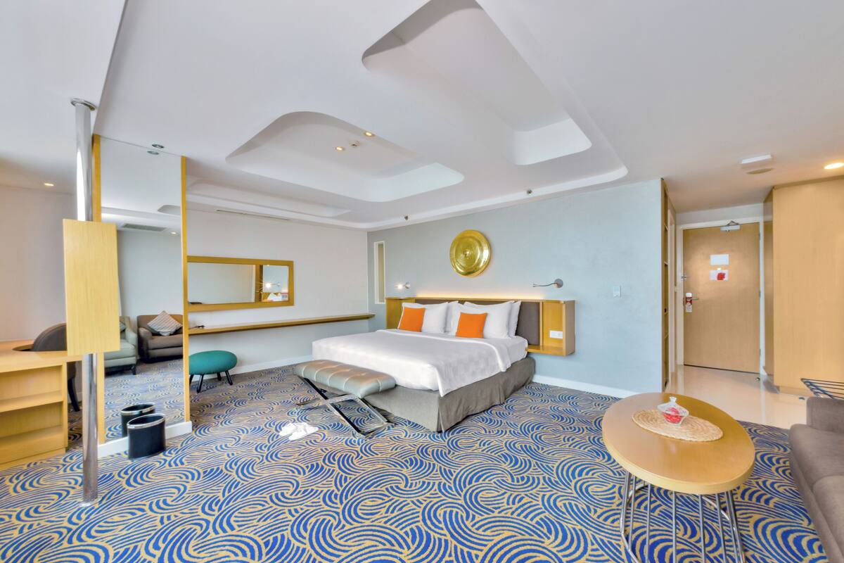 A modern hotel room with a large bed, a desk, a chair, a round table, and a mirror.
