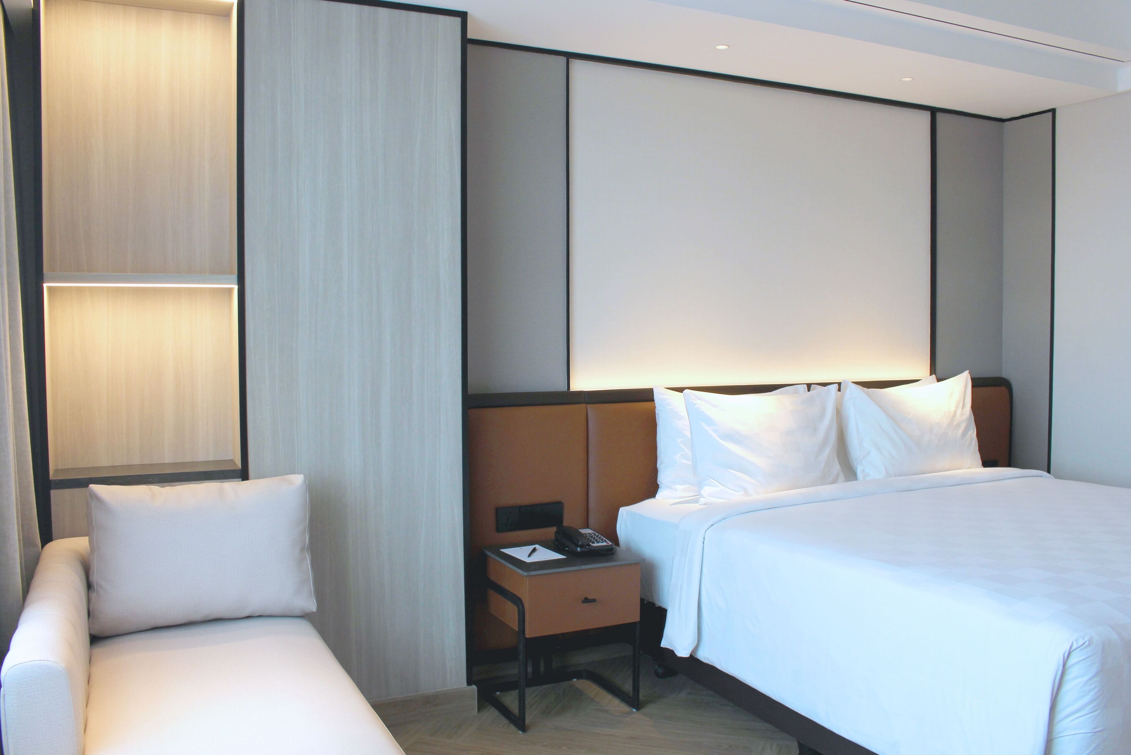 A hotel room with a bed, a nightstand, a chair, and a wall with built-in lighting.