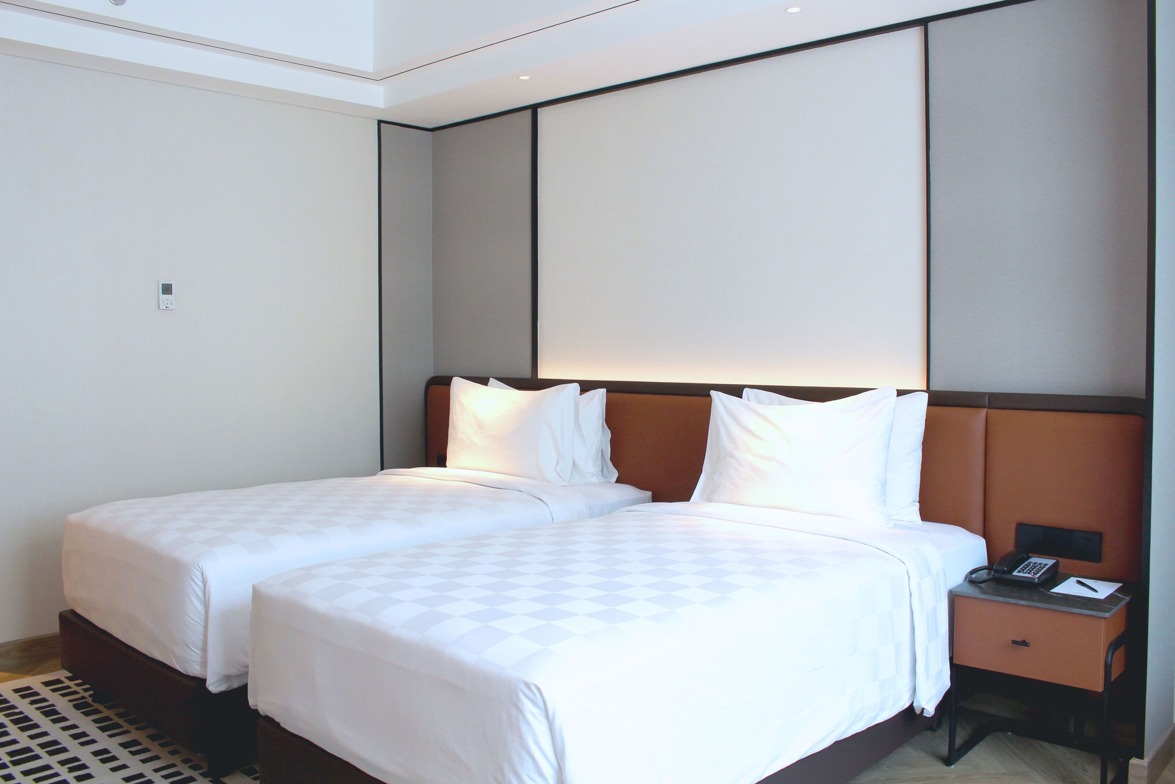 A hotel room with two beds, a nightstand with a phone and notepad, and a wall-mounted panel.