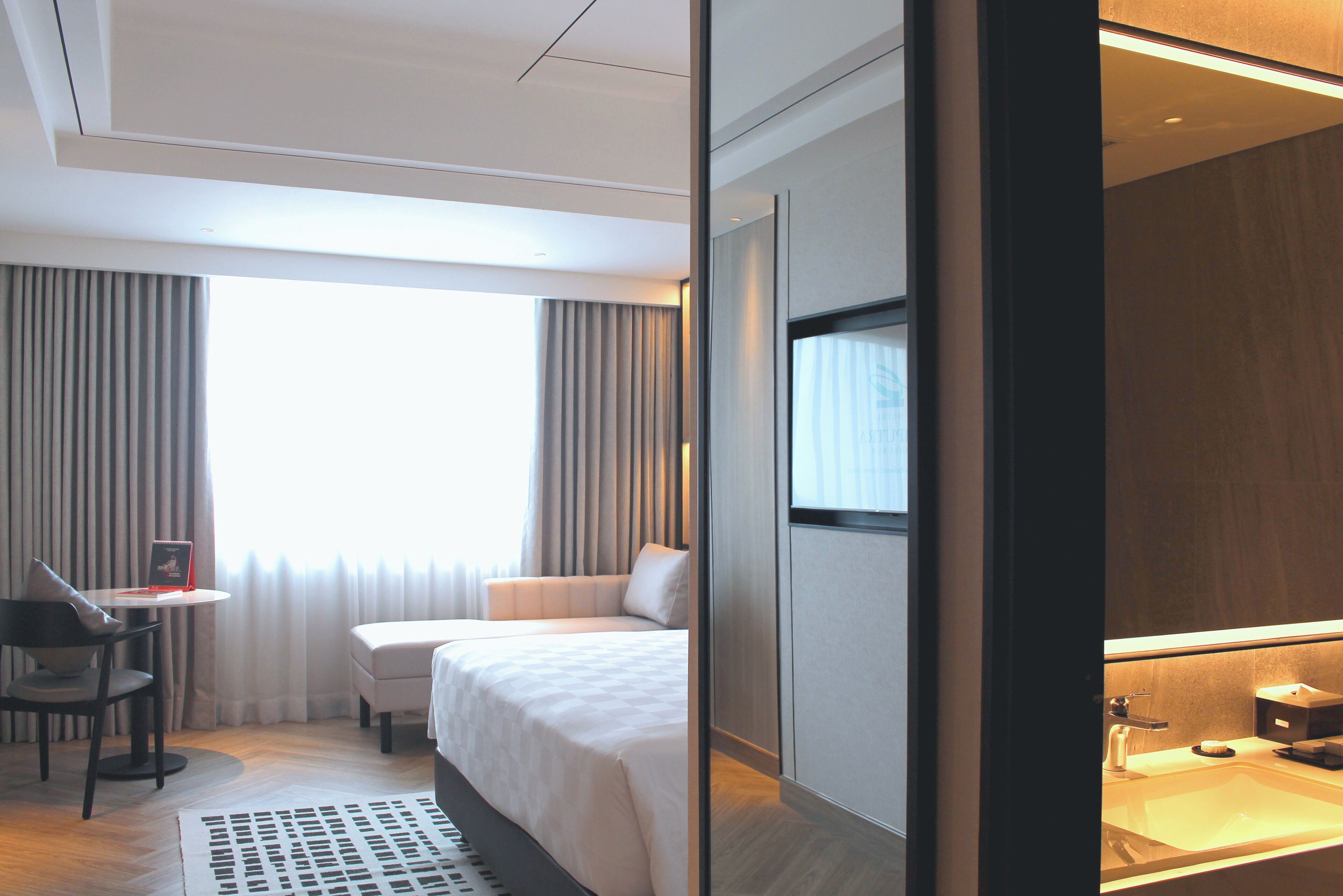 Una camera d'hotel con un letto, una scrivania, una sedia, una finestra con tende e un bagno con lavandino e specchio.