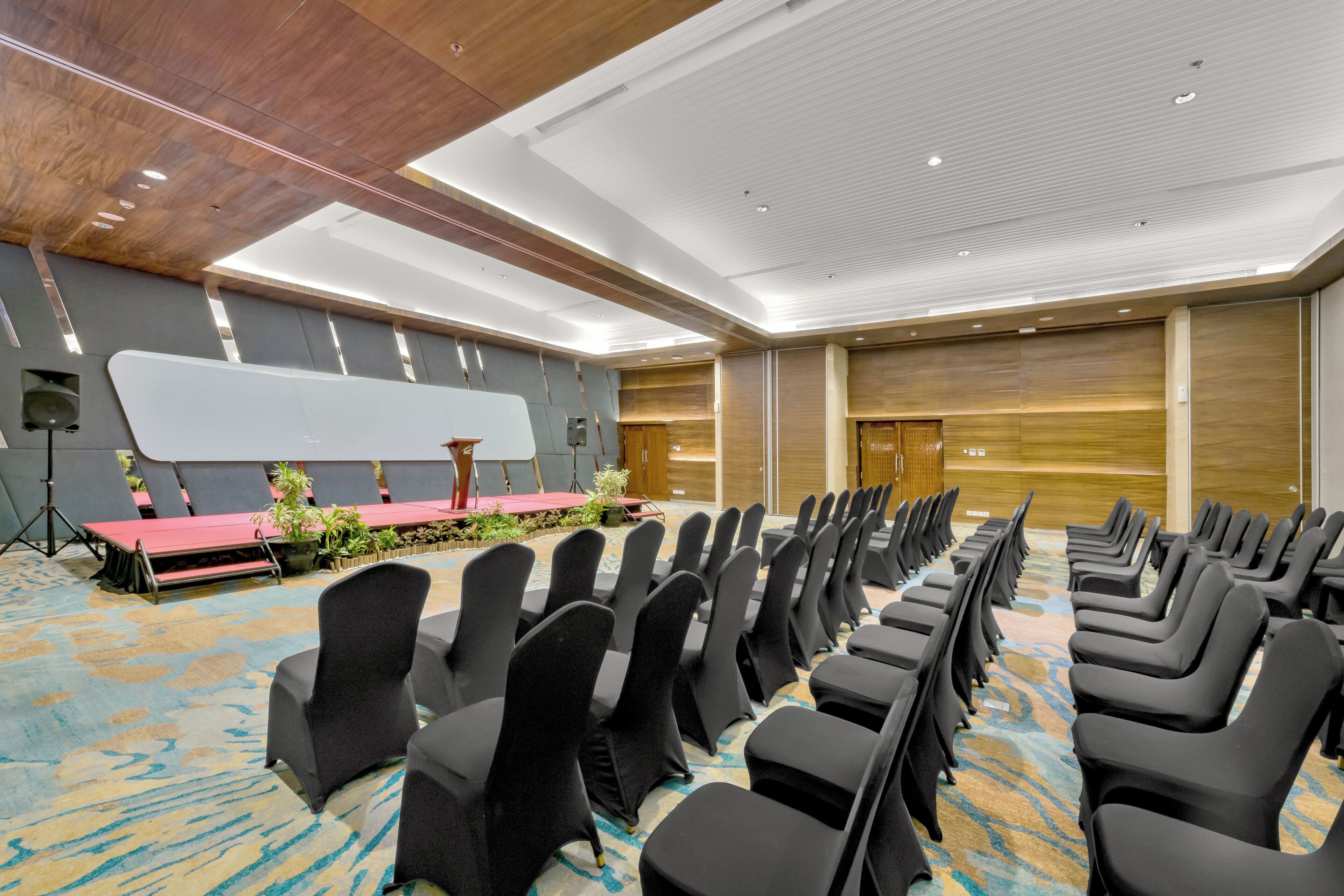 A conference room with rows of chairs, a stage with a red carpet, and a wooden panel wall.