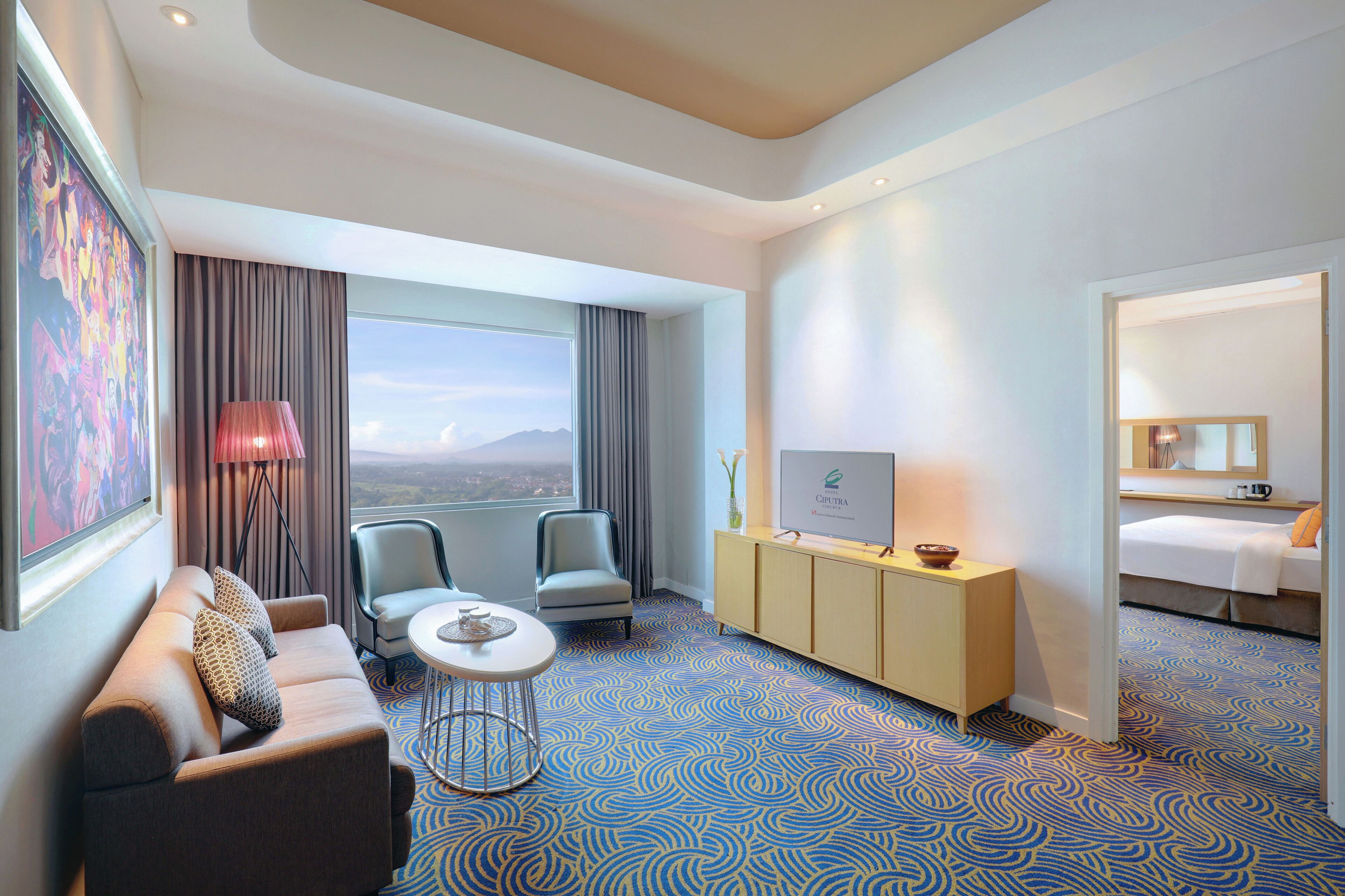 A hotel room with a sofa, two chairs, a small table, a cabinet, a bed, a large window with curtains, and a painting on the wall.