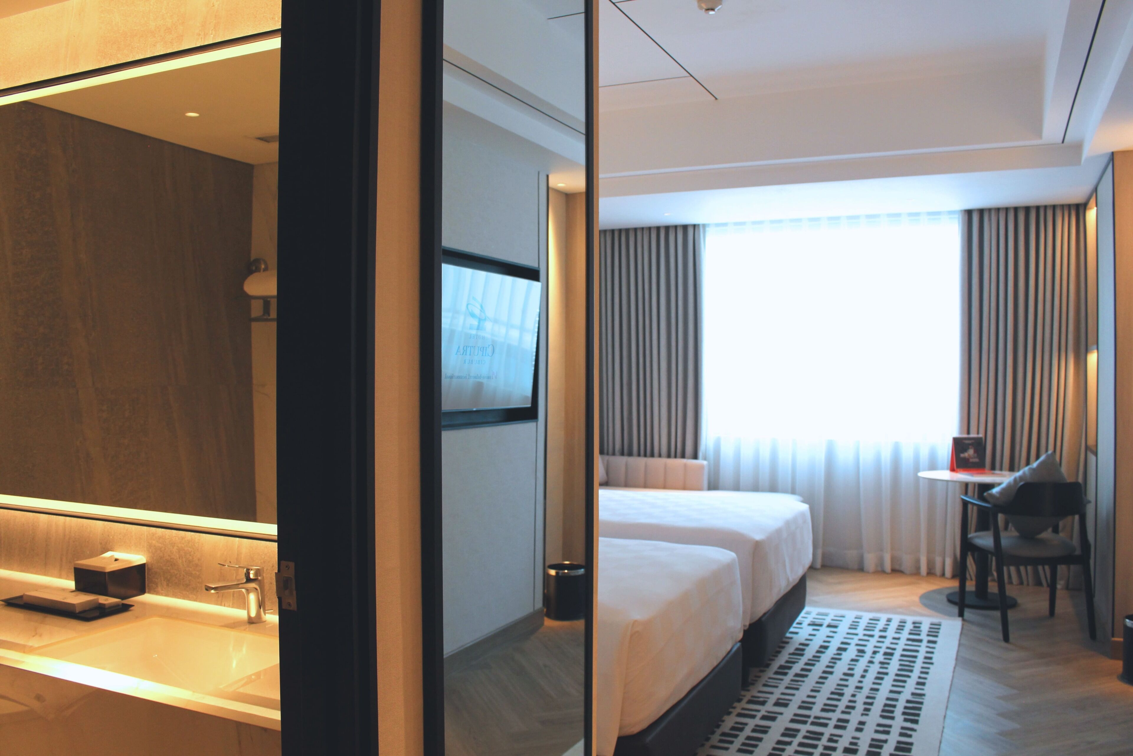 A hotel room with a large mirror, a bed, a desk, and a window with curtains.