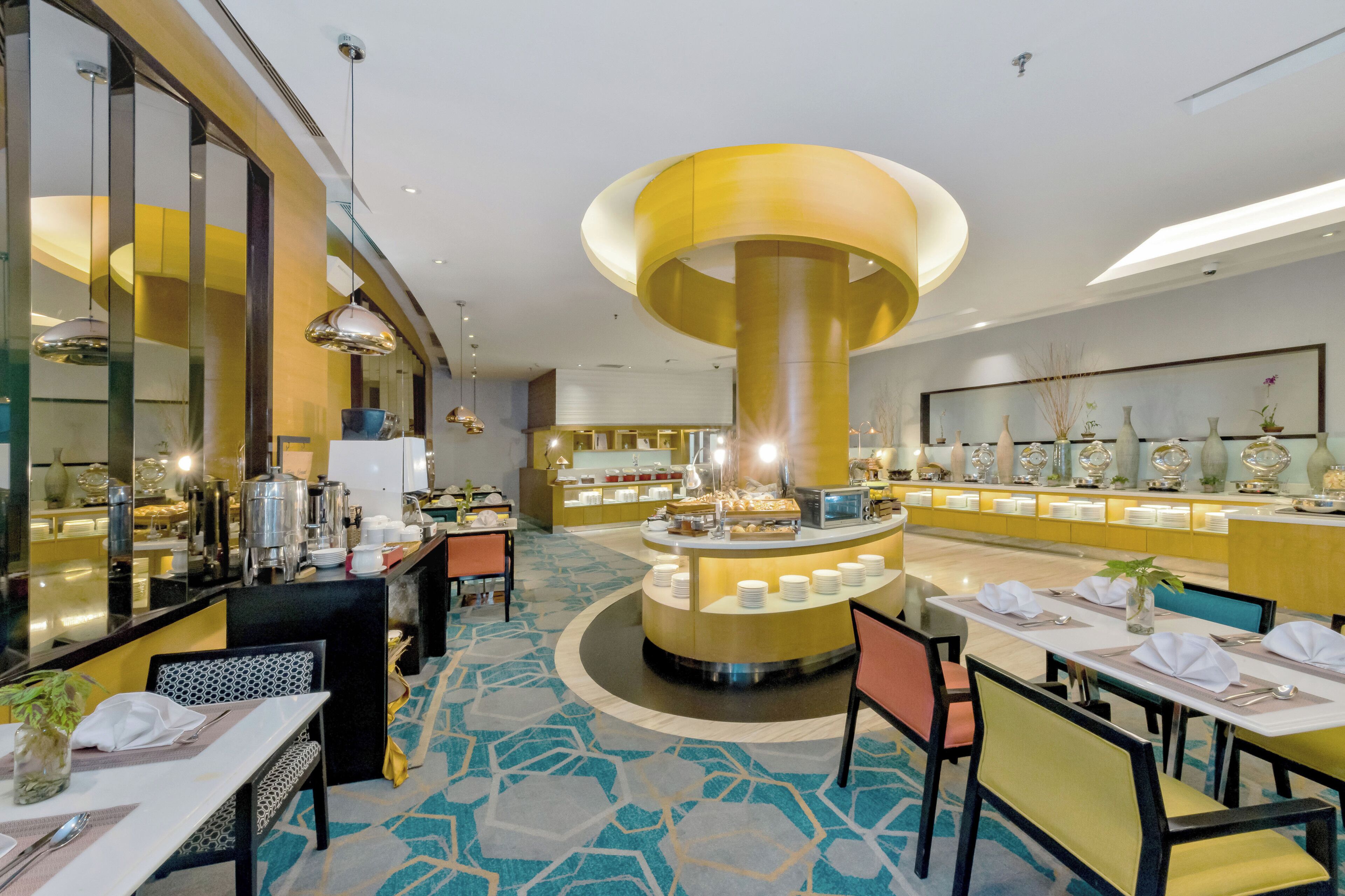 A hotel dining area with a central buffet, round tables, and chairs.