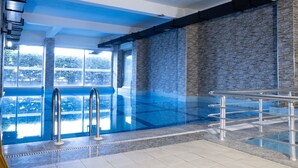 En inomhuspool med väggar av genomskinligt glas, metallräcken och en vägg med steninspirerad yta.