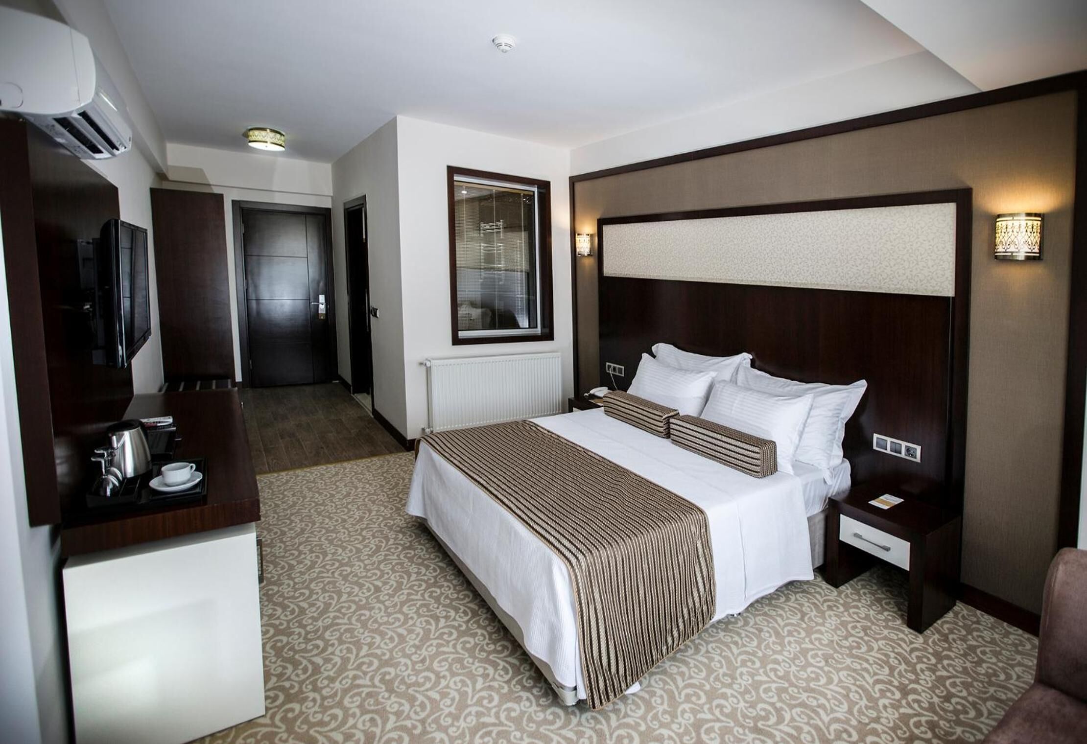 Quarto de hotel com uma cama grande, mesas de cabeceira, uma TV, uma escrivaninha com chaleira e uma janela.