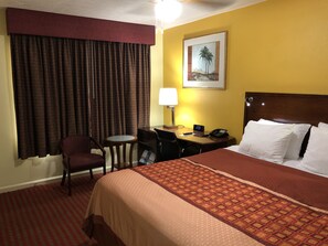 A hotel room with a bed, a desk, a chair, a lamp, and a painting on the wall.